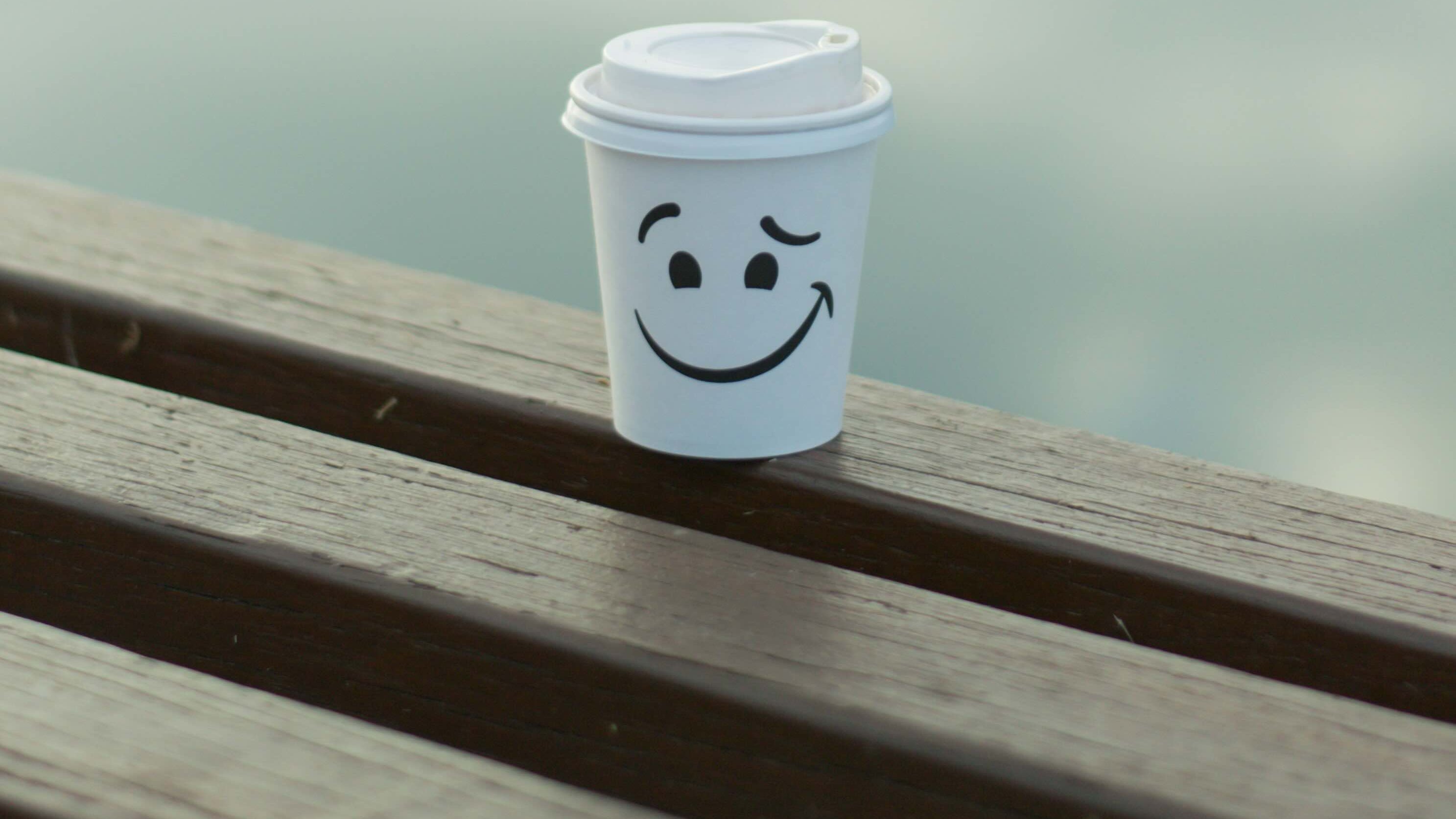 Gobelet de café jetable avec un sourire dessiné au marqueur, symbole d'un masque de bonne humeur malgré la fatigue. Il est 8h, une nouvelle journée commence.