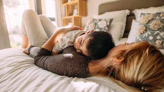 Enfant endormi, allongé sur sa maman — tous deux apaisés sur un lit. Illustration d'un besoin de présence parentale pour s'endormir, avant l'accompagnement sommeil.