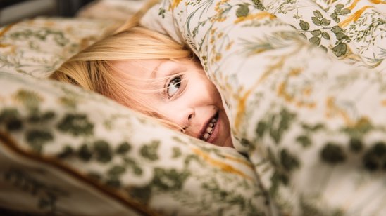 Enfant blond malicieux avec la couette sur la tête, illustrant les défis du coucher et l'accompagnement pour des soirées plus sereines en famille