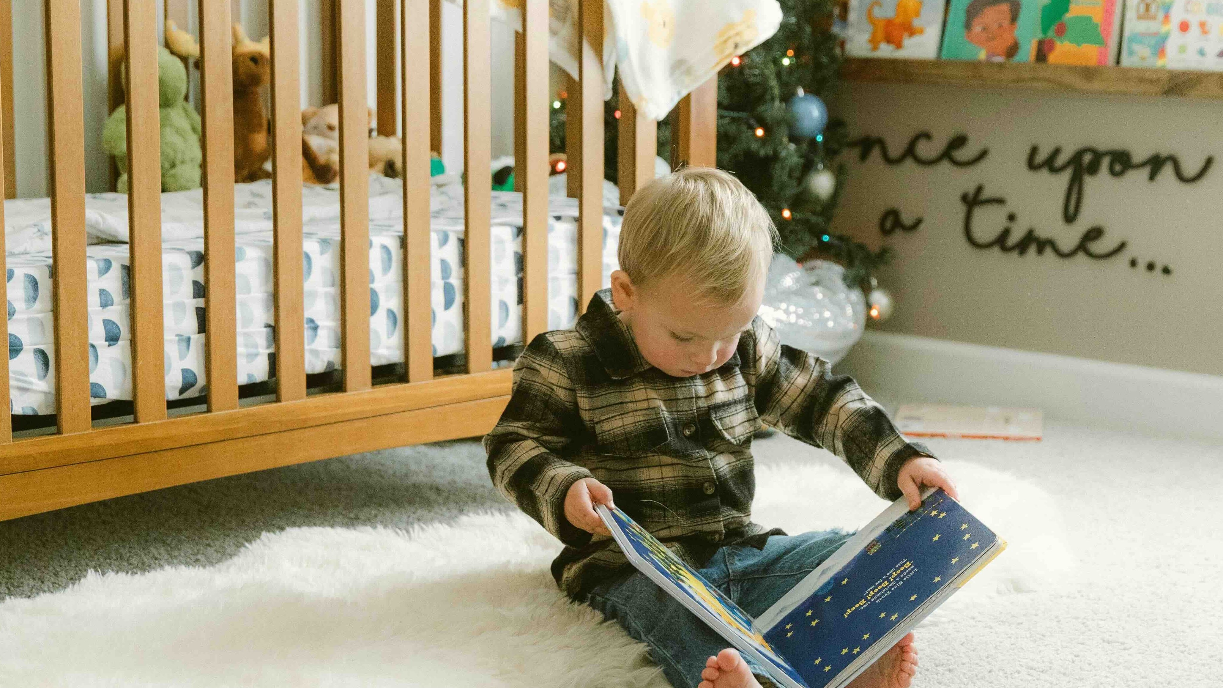 Jeune enfant concentré en train de lire un livre devant son lit à barreaux, dans une chambre paisible avec une bibliothèque adaptée – une scène douce qui évoque les routines du coucher et les moments de calme dans la chambre.