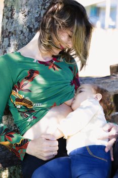 Maman souriante qui allaite son bébé adossée à un arbre – image de sérénité retrouvée après des nuits difficiles, grâce à un accompagnement sommeil respectueux de l'allaitement, comme pour Émeline et son bébé Milo.