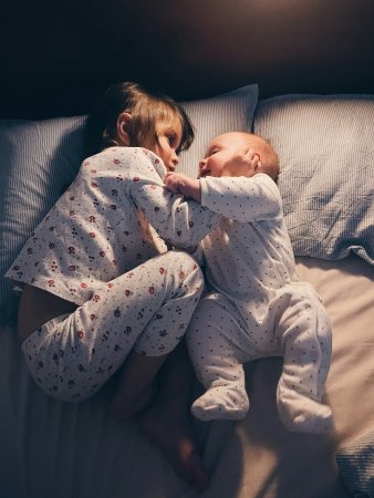 Bébé et sa grande sœur allongés face à face dans un lit, calme et complices, lumière douce de la nuit, ambiance apaisante pour le sommeil.
